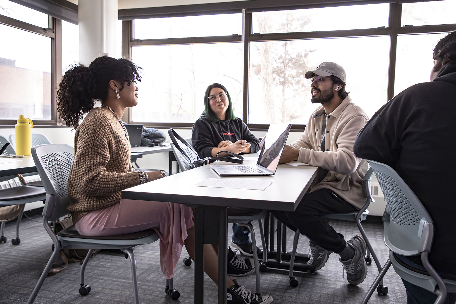 Three college students sit around a white table in a classroom with large windows, working on laptops and talking.