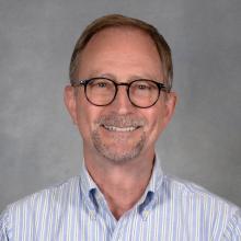 Man with glasses, short brown hair and close-cropped beard wearing striped blue button-down shirt