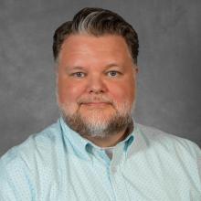 Man with short dark hair and beard wearing light blue shirt