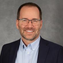 Man with glasses, short brown hair and close-cropped beard wearing dark blue jacket and light blue button-down shirt