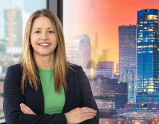 Woman with brown hair wearing a green shirt and blue jacket. Denver skyline in the background.